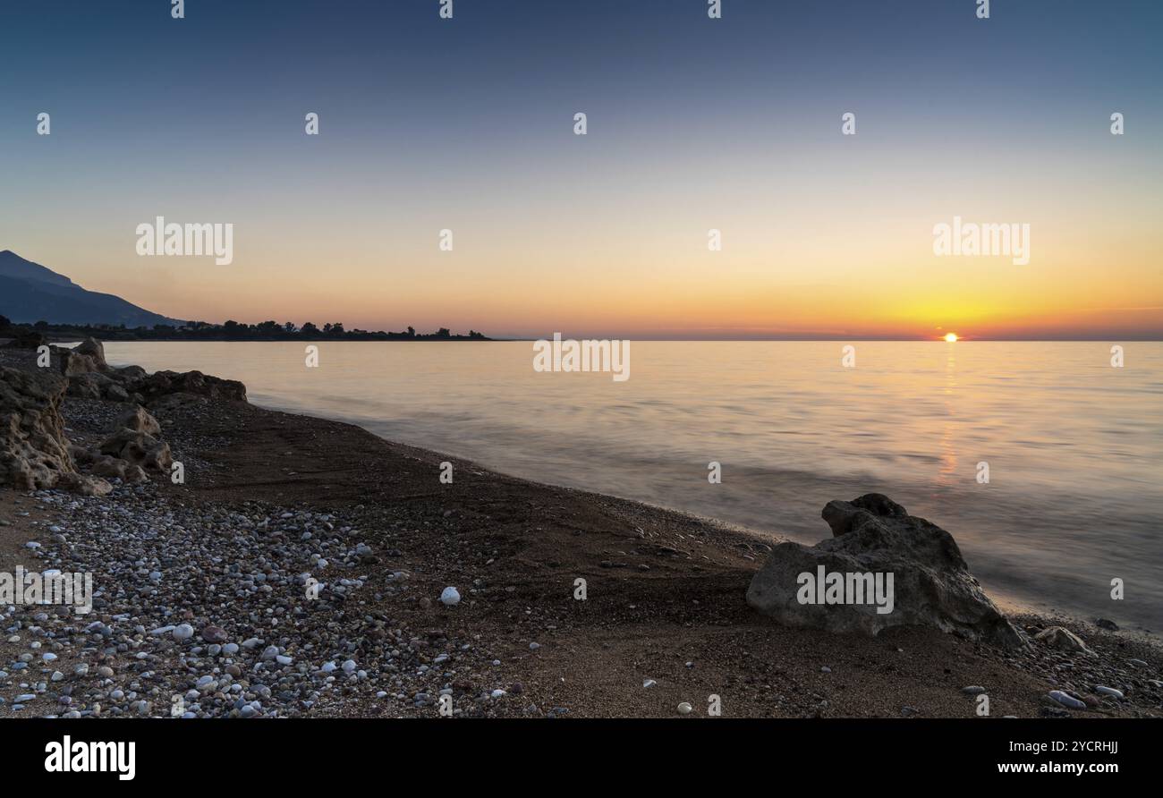 Un tramonto tranquillo sull'oceano con una silhouette di montagne e una spiaggia di ciottoli con massi di roccia in primo piano Foto Stock