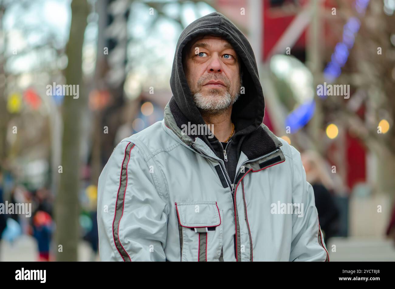 Ritratto di strada di un uomo anziano di 50-55 anni che indossa una felpa con cappuccio sulla testa su uno sfondo urbano sfocato. Foto Stock