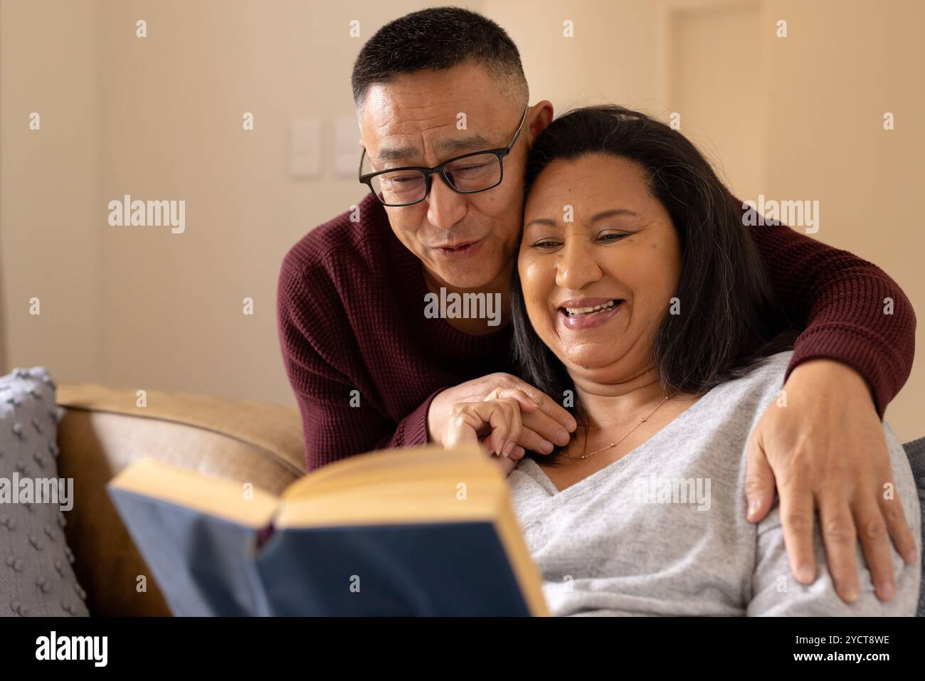 Una coppia senior multirazziale che legge insieme su un comodo divano, condividendo momenti di gioia, a casa Foto Stock