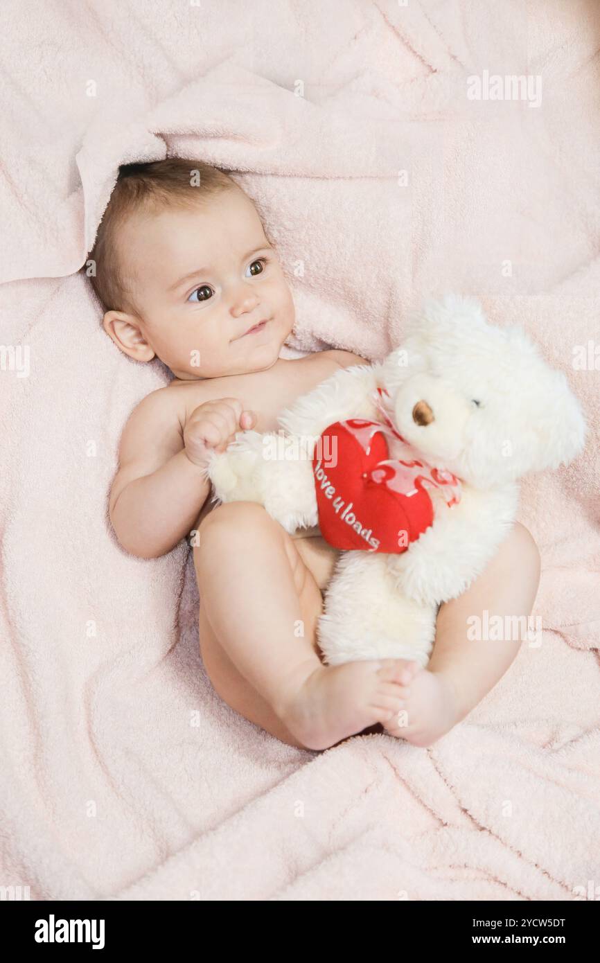 Un bambino adorabile e carino che tiene allegramente in braccio un orsacchiotto mentre si sdraia su una morbida coperta rosa, mostrando pura innocenza Foto Stock