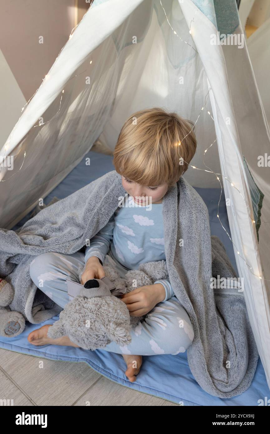 un bambino carino in pigiama che parla con un orsacchiotto seduto in pigiama in un wigwam. fantasy, amico immaginario, giocare con giocattoli morbidi, la gioia di chil Foto Stock