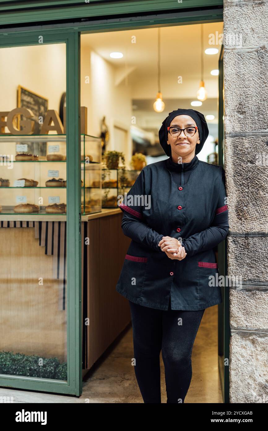 Un fiducioso pasticcere vegano si erge fuori dalla sua panetteria, sorridendo verso la macchina fotografica, vestito con un'uniforme da chef professionista, contro l'invitante Foto Stock