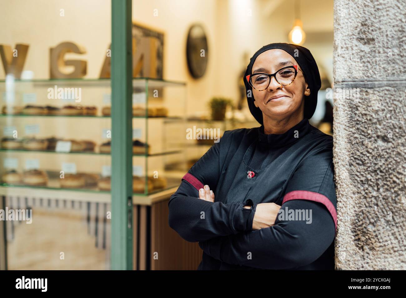Donna sorridente, in piedi con fiducia nella sua panetteria vegana con ciambelle visibili sullo sfondo, rappresenta il successo e un imprenditore attento alla salute Foto Stock
