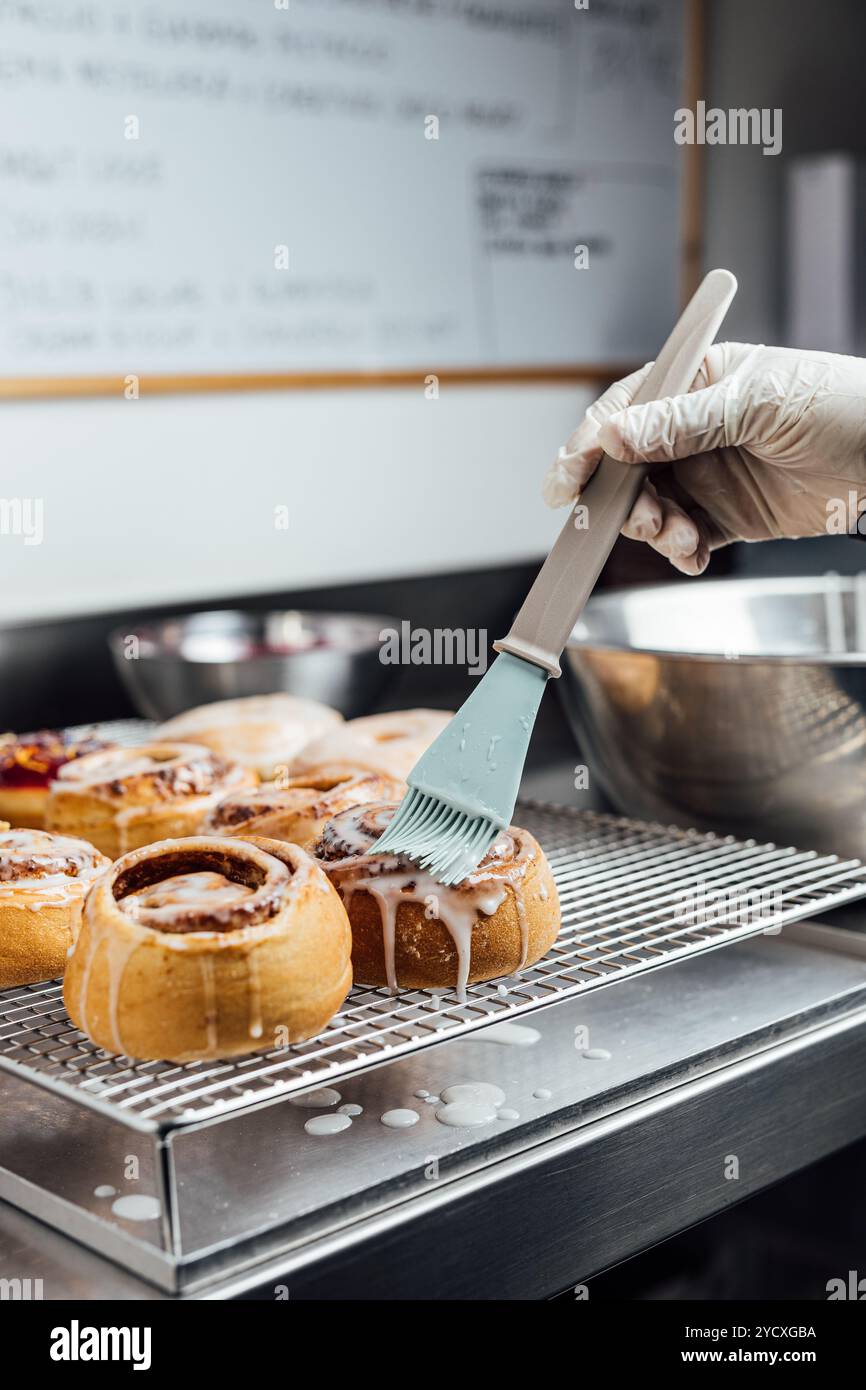 Un pasticcere vegano decora meticolosamente un lotto di dolci alla cannella appena sfornati con glassa in una panetteria insieme a diverse ciambelle smaltate, mostrando il cu Foto Stock