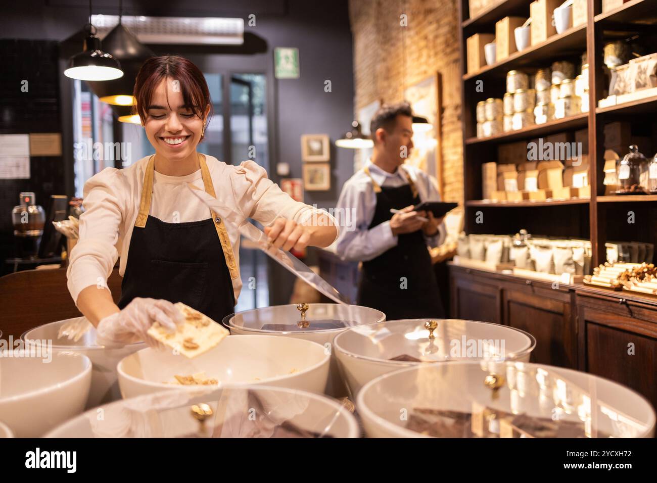 Una donna sorridente che mette il cioccolato in un negozio di cioccolato di prima qualità, accompagnata da un collega che gestisce i prodotti in background Foto Stock