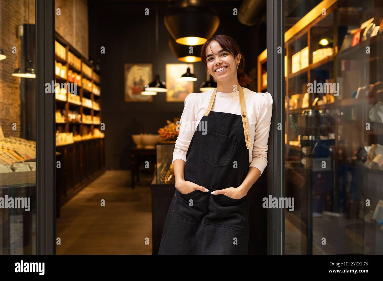 Una giovane donna allegra che indossa un grembiule si trova all'ingresso di un'accogliente e ben illuminata cioccolateria, circondata da scaffali riforniti di varie cioccolate Foto Stock