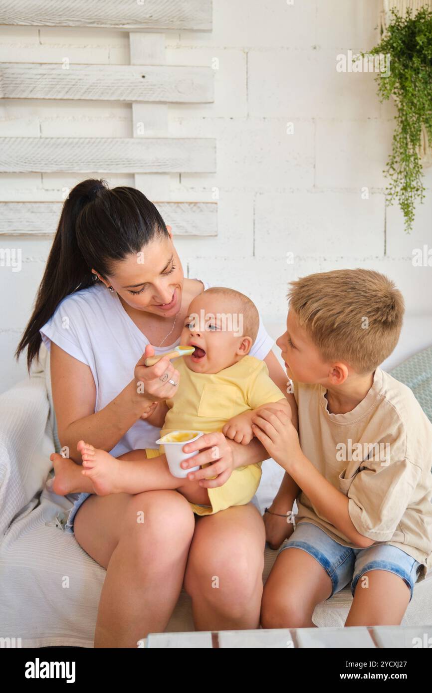 Un momento di tenerezza mentre una madre, seduta comodamente, nutre il bambino con un cucchiaio, mentre il bambino più grande guarda attentamente questa scena mozzafiato Foto Stock