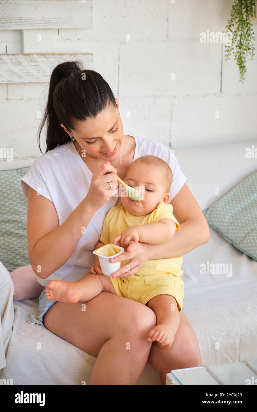 Un momento tenero, catturato da una madre che nutre delicatamente il bambino con un cucchiaio, entrambi comodamente seduti su una panchina bianca a casa, l'impostazione è più luminosa Foto Stock