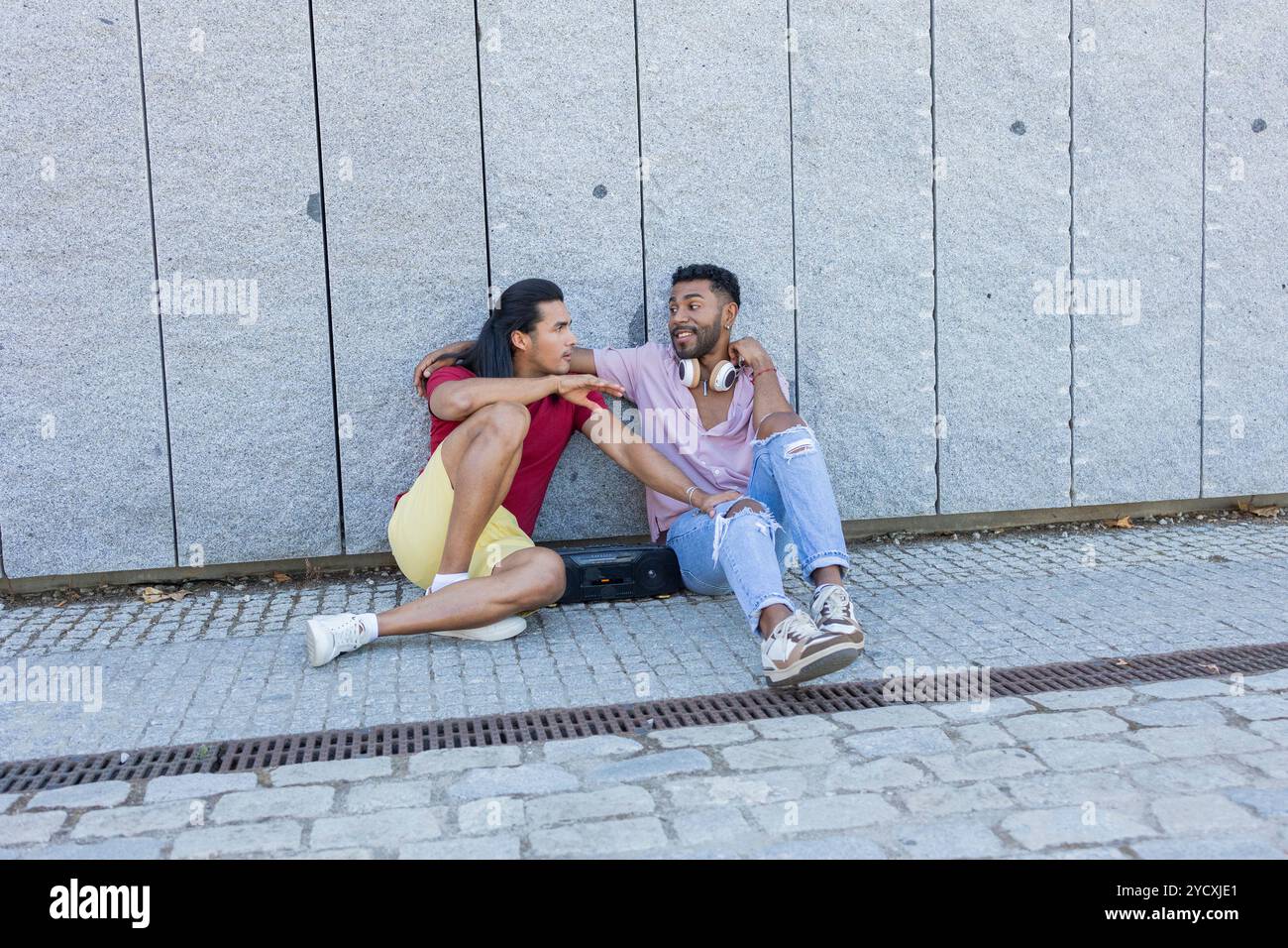 La coppia gay condivide un momento di gioia seduto contro un muro in una giornata di sole, che riflette le diverse culture dell'America Latina e dell'afroamericano heritag Foto Stock
