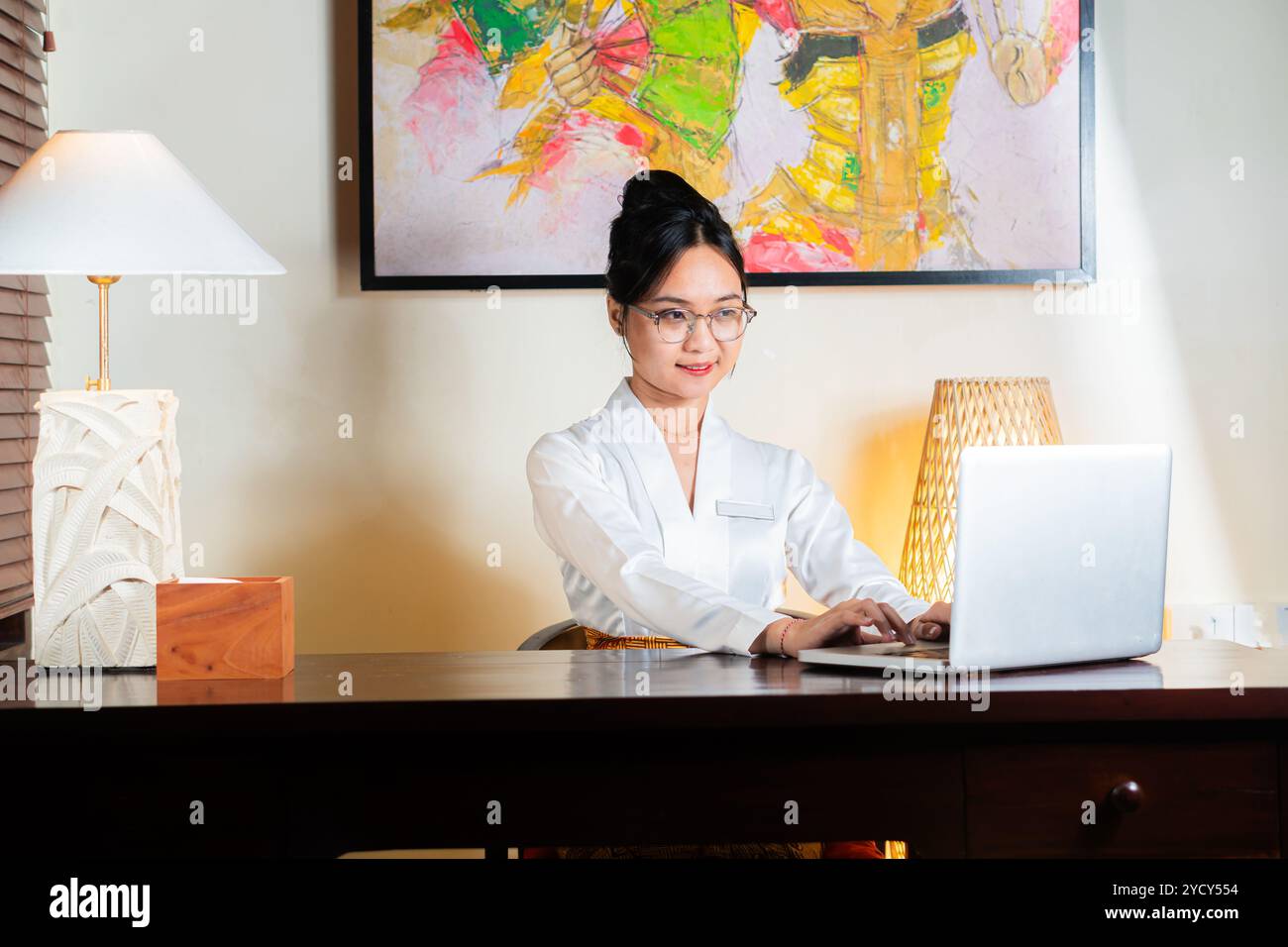 Una donna con una camicetta bianca che lavora su un computer portatile a una scrivania in una stanza ben illuminata con un dipinto colorato sulla parete. Foto Stock