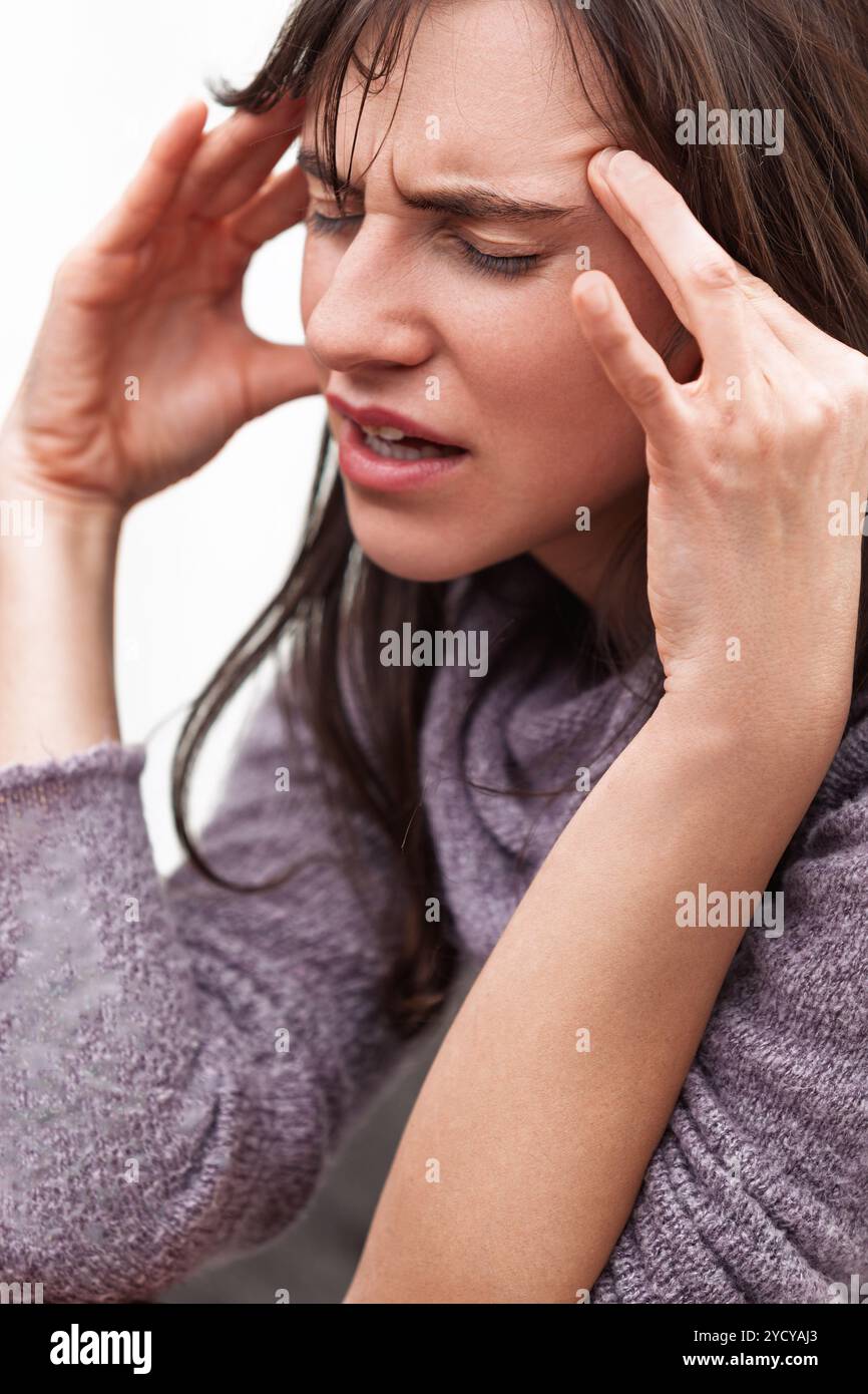 Giovane donna che tocca i suoi templi, cercando di alleviare un mal di testa di tensione con un'espressione dolorosa, mostrando segni di stress e sofferenza a casa Foto Stock