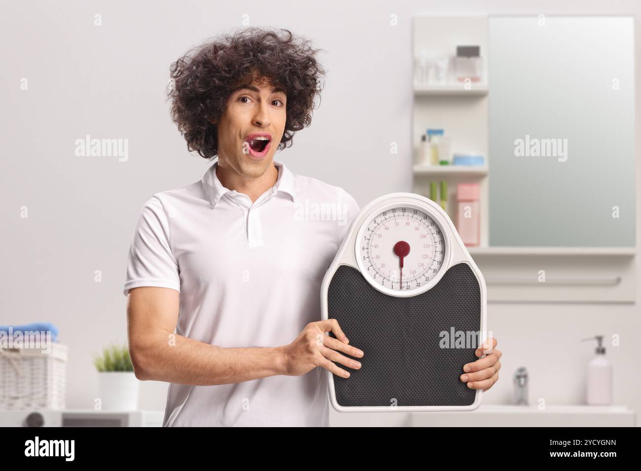 Giovane uomo con capelli ricci che tiene una bilancia in bagno Foto Stock