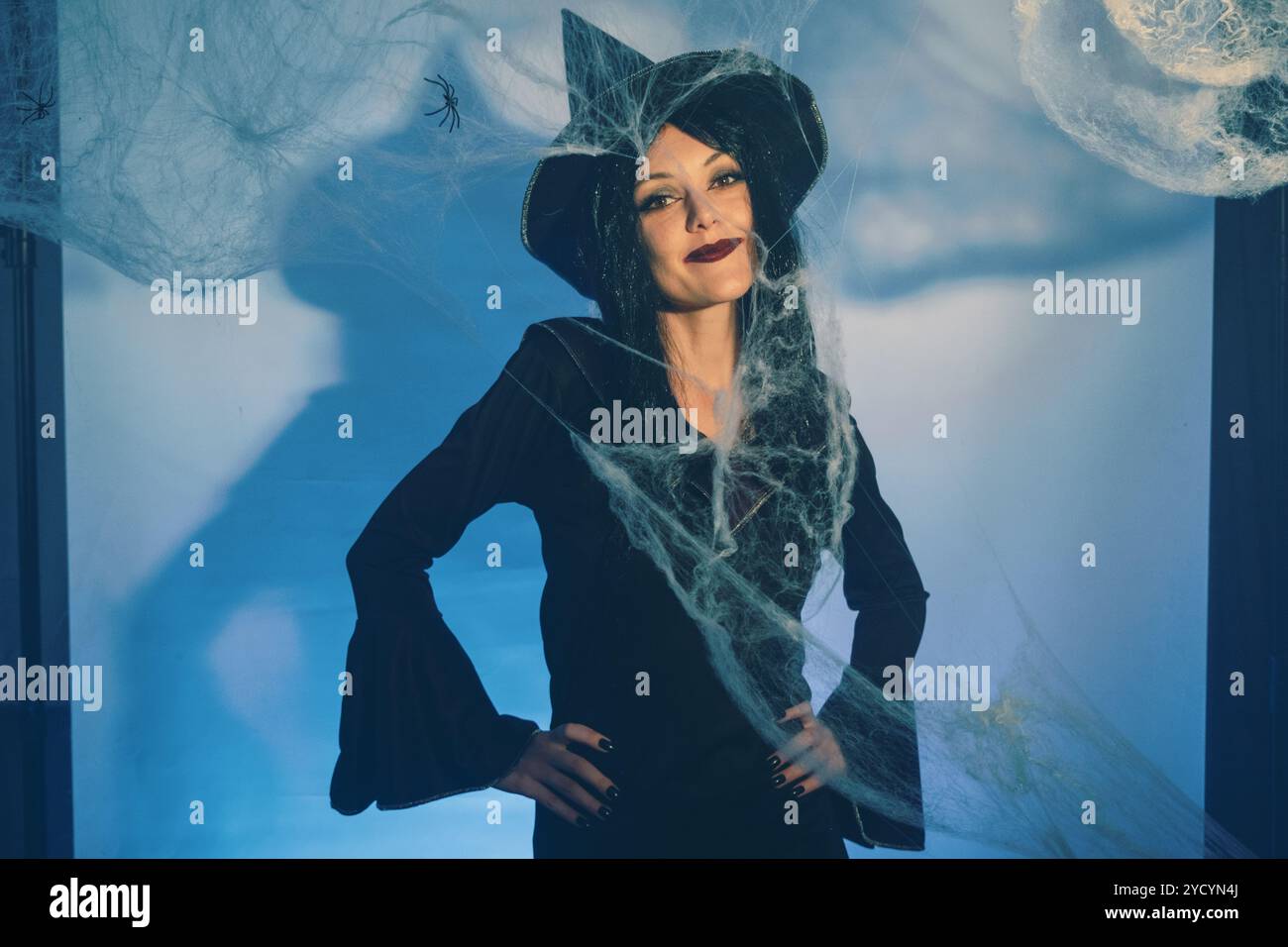 Una strega affascinante posa davanti alle ragnatele, le sue mani alzate con un gesto giocoso. Indossa un cappello appuntito e un sorriso sicuro, creando capricci Foto Stock