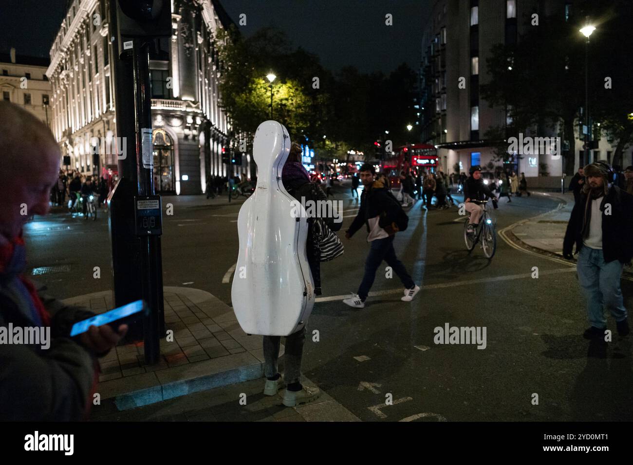 Una musicista classica cammina attraverso l'attraversamento pedonale dello Strand portando con sé il suo strumento per violoncello di notte in IT's Case, Londra, Inghilterra, Regno Unito Foto Stock