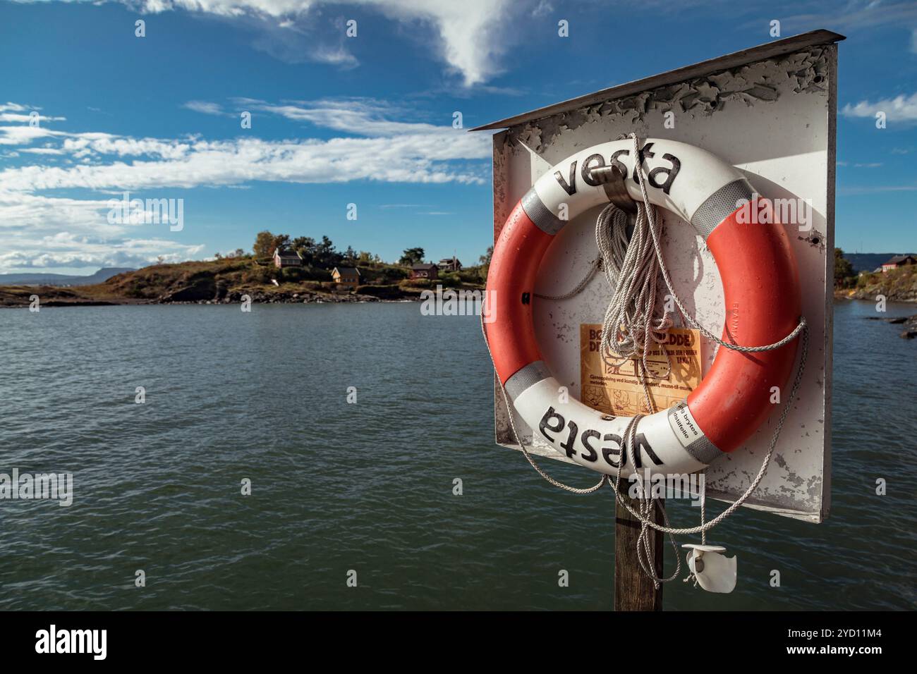 Un salvagente arancione luminoso è appeso su un palo vicino all'acqua, con l'isola Gressholmen visibile in lontananza sotto un cielo limpido. La scenografia incarna recreatio Foto Stock