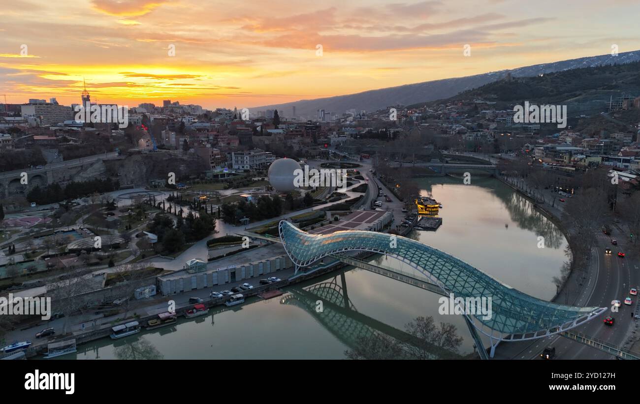 L'iconico Ponte della Pace attraversa il fiume Kura al tramonto a Tbilisi, Georgia, con la sua struttura in vetro e acciaio illuminata contro un cielo dorato. Foto Stock