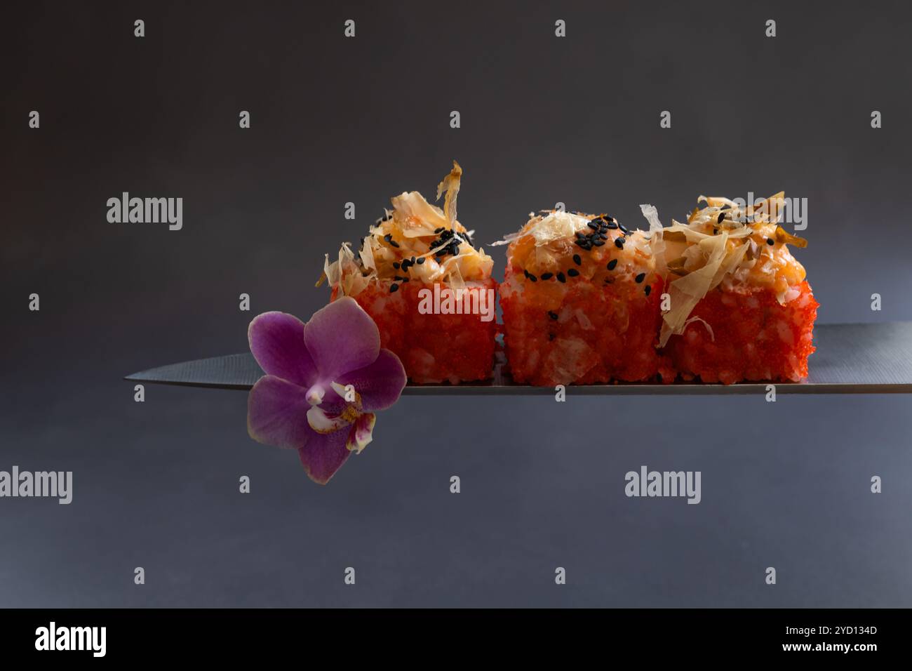 Eleganti sushi Rolls serviti su una lama decorata con un fiore d'orchidea su sfondo scuro Foto Stock