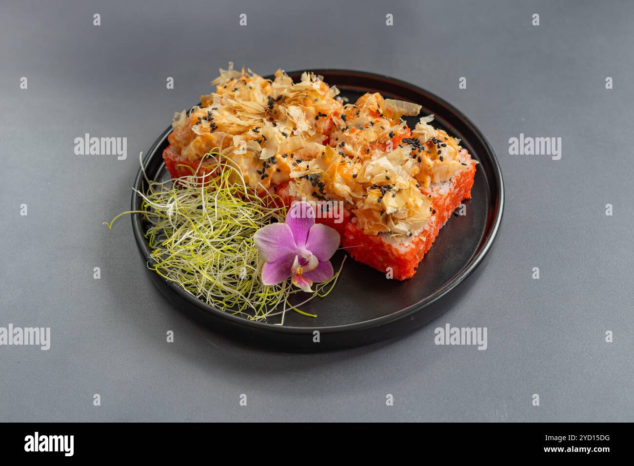 Deliziosi sushi Rolls serviti su un piatto nero con microgreen e un fiore di orchidea su sfondo grigio Foto Stock