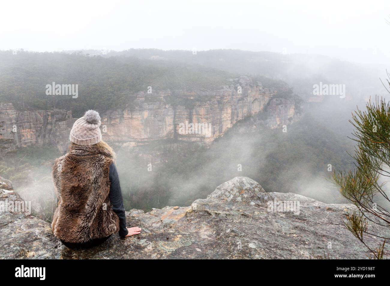 Donna seduta sulla sporgenza della scogliera in cima alla montagna che guarda fuori nella nebbia nebbiosa Foto Stock