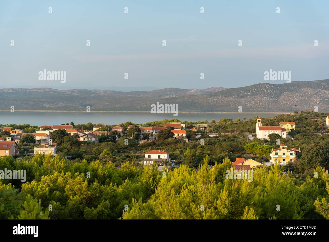 Affascinante villaggio rurale immerso nel verde con montagne lontane e un lago tranquillo sotto un cielo morbido e nuvoloso. Foto Stock