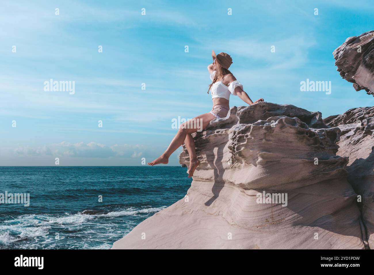 Donna che si gode il sole estivo, seduta accanto all'oceano e indossa un cappello Foto Stock