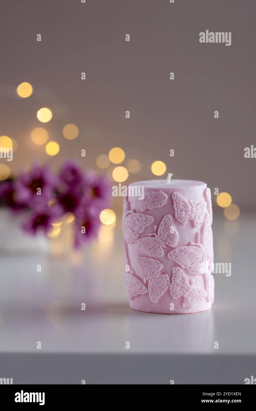 Candela decorativa rosa con motivo a farfalla. Fiori e luci sullo sfondo con spazio di copia. Arredi per la casa. Interni accoglienti. Preparazione di candele di cera di soia. Foto Stock