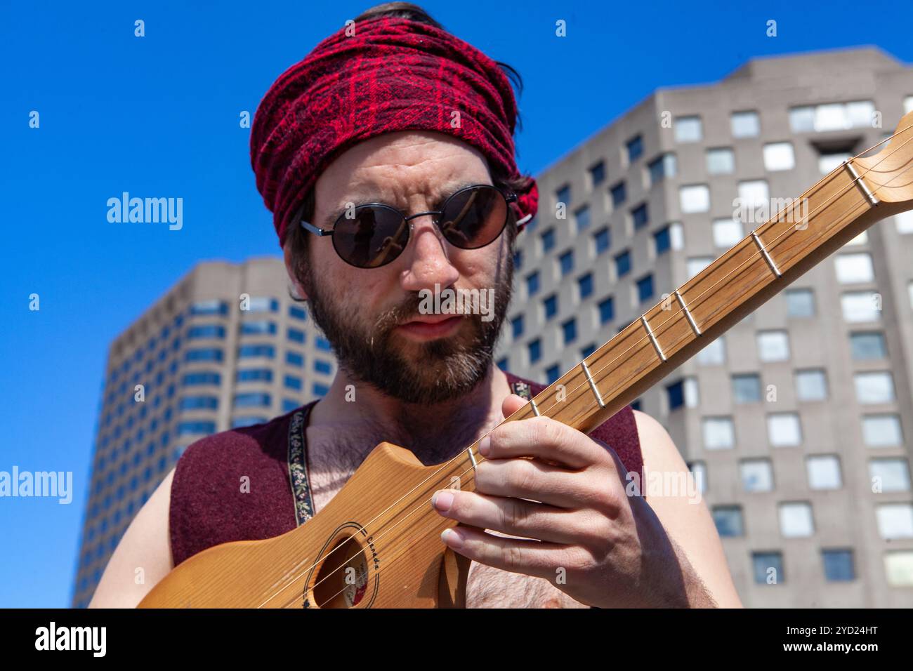 Un primo piano ritratto di un sacro con red bandana uomo tenendo un piccolo acustico chitarra merlin in città, corporate grattacielo in background downtown Foto Stock