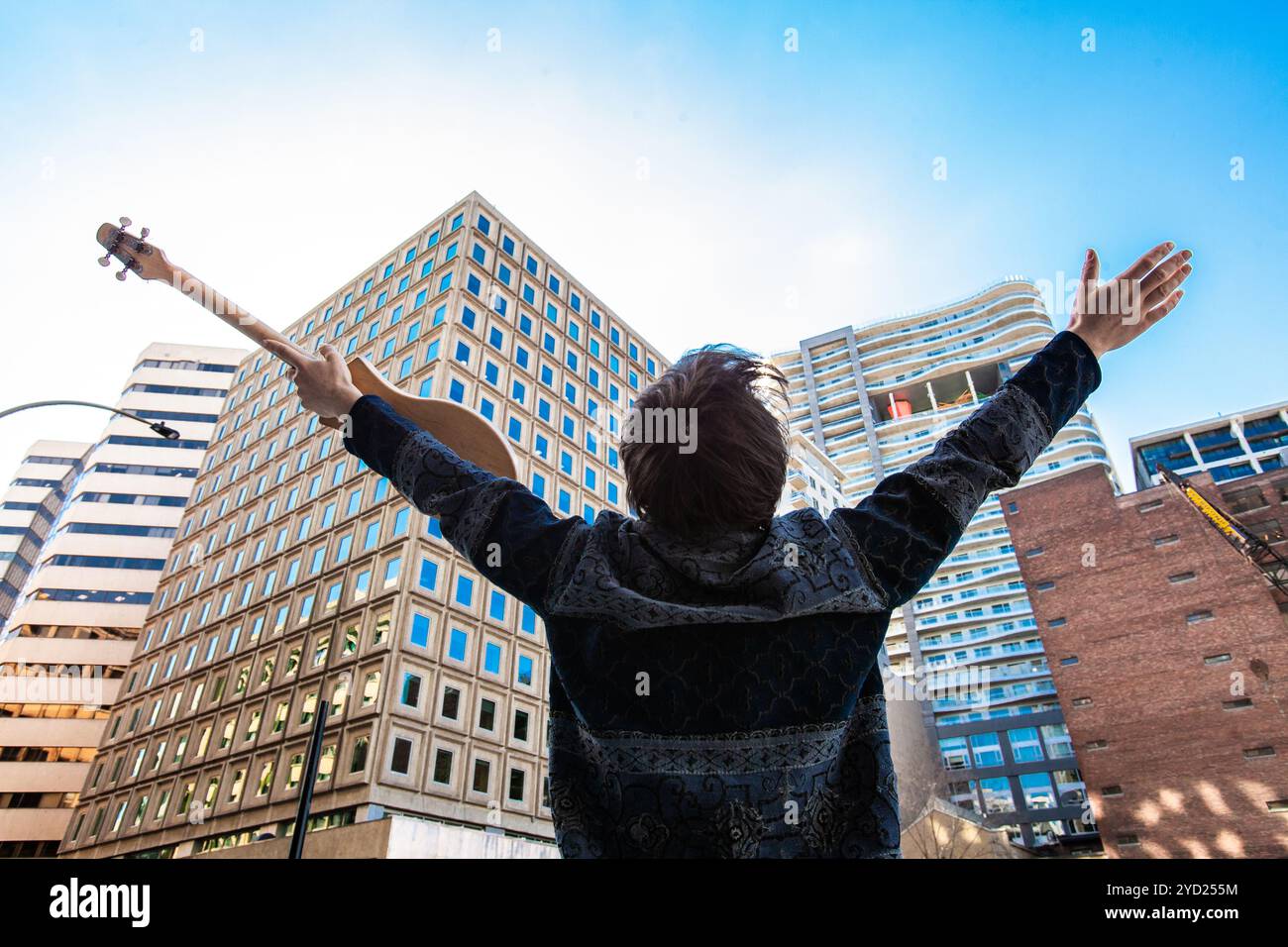 Un basso angolo e la vista posteriore di un uomo felice tenendo un merlin guitar, centro permanente con i bracci sollevati, ispirato dalla città moderna architettura. Foto Stock