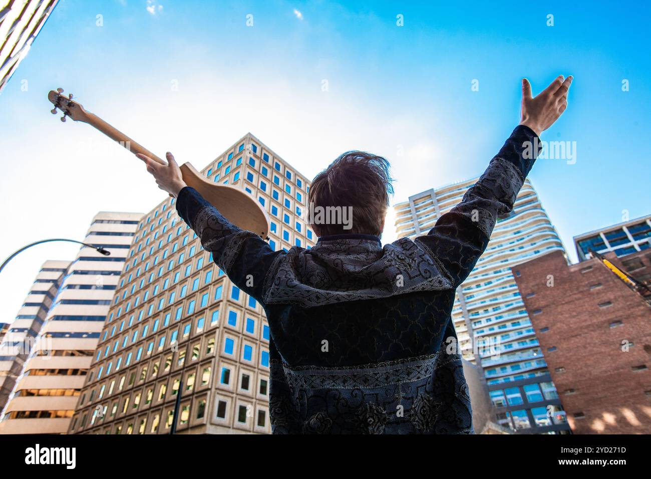 Un uomo di boemia tenendo una piccola chitarra acustica è visto dal retro, tenendo le braccia in alto verso la costruzione di edifici commerciali in centro città. Foto Stock