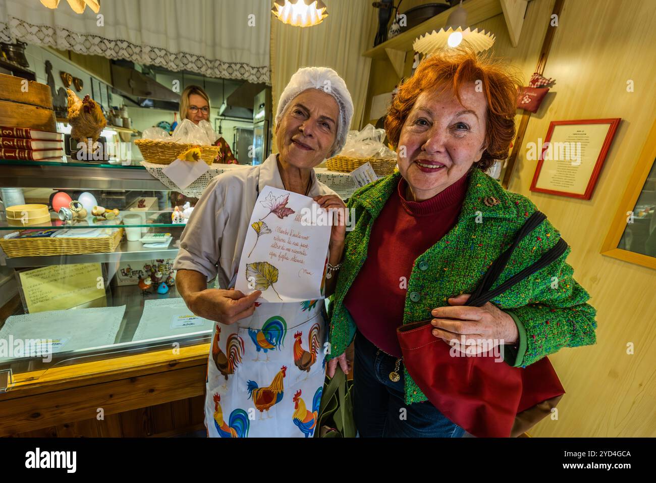 Un dipendente della pastaia Le Sfogline mostra a un cliente una carta manoscritta illustrata con un motivo a foglia. In secondo piano, un secondo dipendente è visibile in cucina, riflesso nello specchio del negozio. Via Belvedere, Bologna, Emilia-Romagna, Italia. Foto Stock