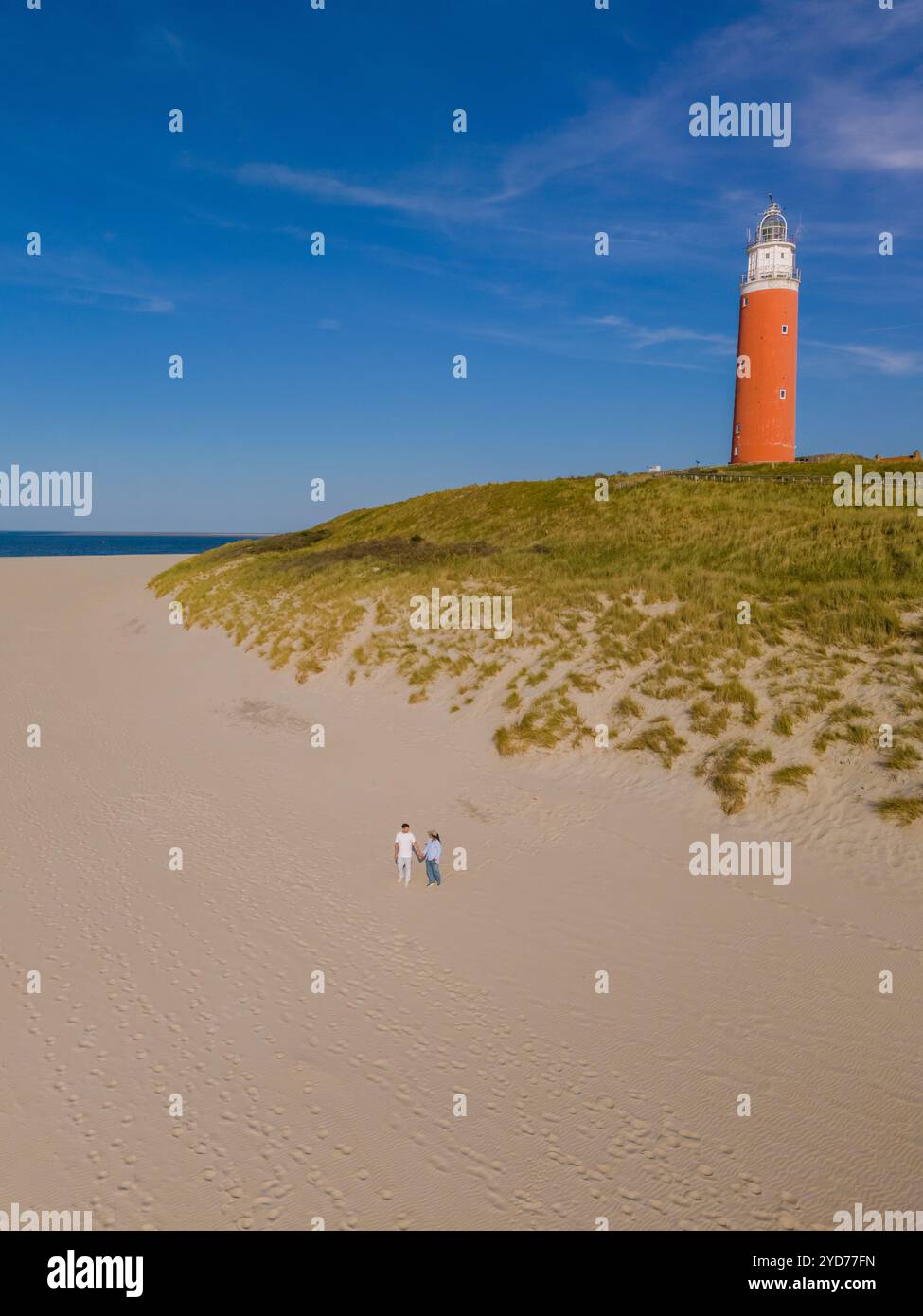 Una coppia si trova fianco a fianco su una spiaggia sabbiosa vicino ad un faro rosso e bianco, bagnata dalla calda luce del tramonto sullo sfondo Foto Stock