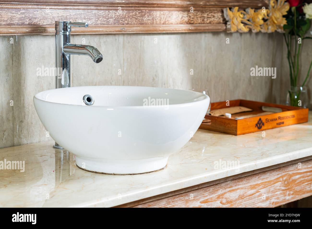 Un moderno lavabo da bagno con un lavabo bianco rotondo e rubinetto cromato su un ripiano in marmo, con un vassoio in legno e fiori decorativi sullo sfondo Foto Stock