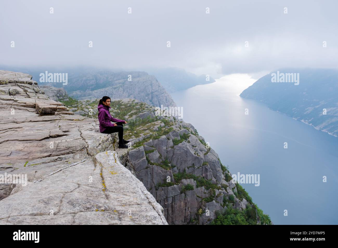 Una donna siede sul bordo della scogliera del Preikestolen in Norvegia, affacciata su uno splendido fiordo. La mattina nebbiosa si aggiunge al dramma Foto Stock