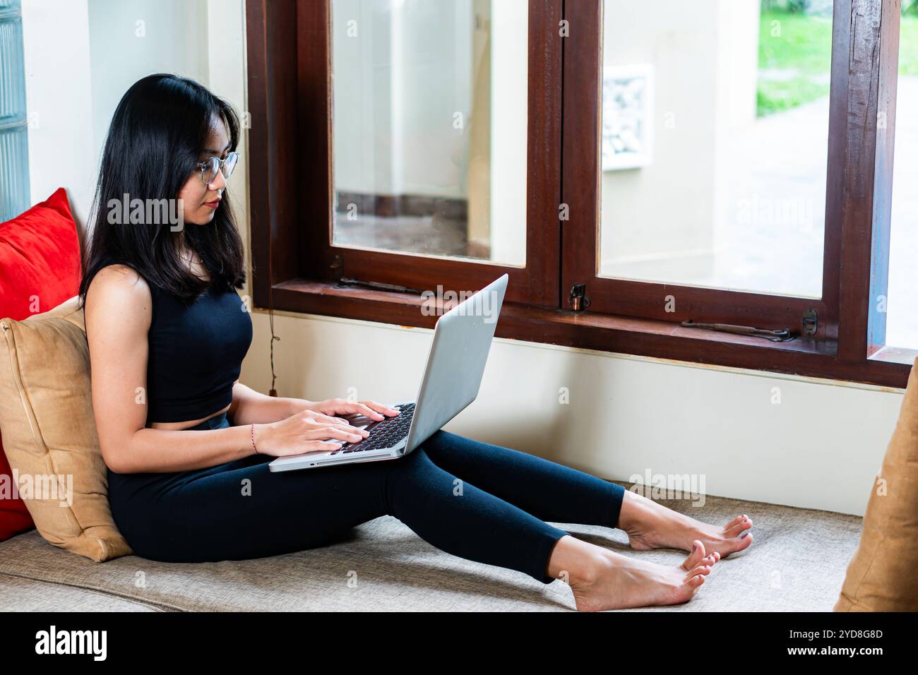 Una giovane donna seduta su un cuscino vicino a una finestra, che lavora su un notebook. Ha lunghi capelli neri e indossa occhiali, vestita con canotta nera e leggin Foto Stock