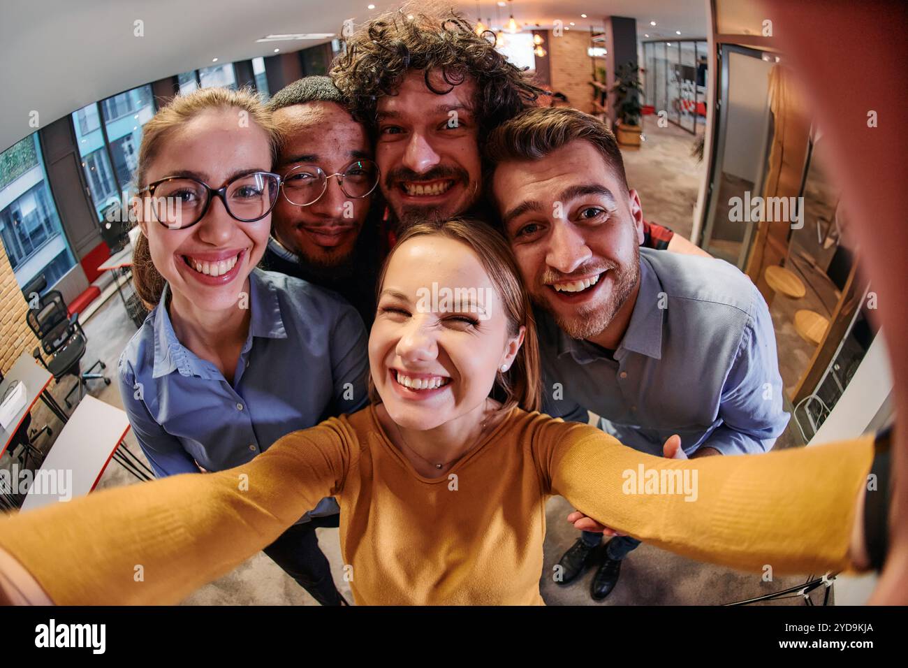 Un gruppo eterogeneo di professionisti si prende una pausa dalle loro attività in un moderno ufficio di startup per scattare un selfie creativo, mostrando il lavoro di squadra a. Foto Stock