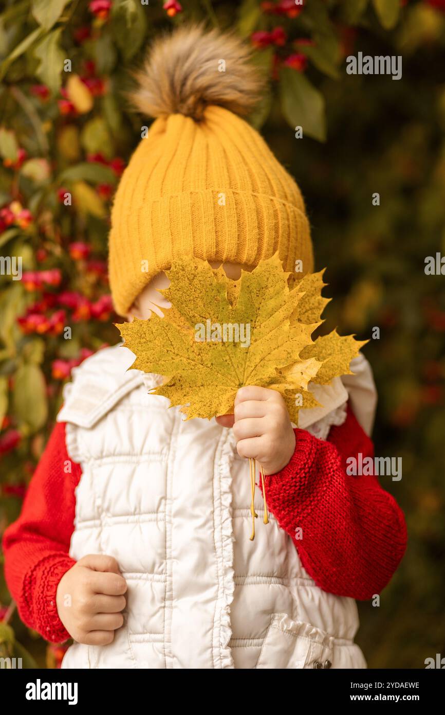 Un bambino che indossa un cappello giallo brillante e un gilet bianco nasconde giocosamente il viso con una grande foglia gialla, circondato da colorate foglie autunnali in una Foto Stock