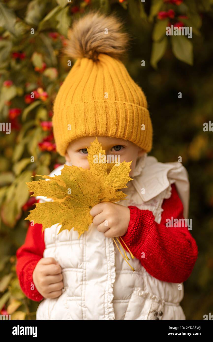 Un bambino allegro vestito con un maglione rosso e un accogliente cappello giallo gode della bellezza dell'autunno tenendo una grande foglia dorata in un giardino colorato. Foto Stock