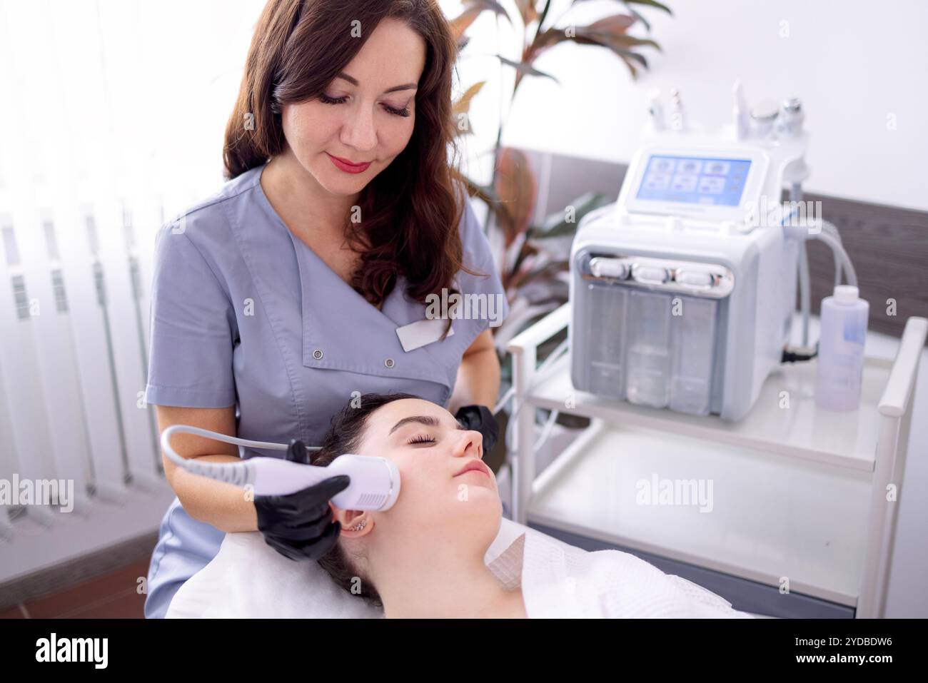 Una giovane donna in un accappatoio mente e riceve una procedura di pulizia del viso utilizzando un dispositivo Foto Stock