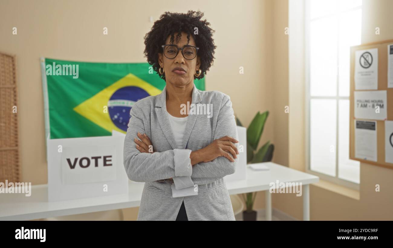 Una bella giovane donna afroamericana con i capelli ricci e le braccia incrociate si trova all'interno di una sala del college elettorale brasiliano con una bandiera e una banconota elettorale Foto Stock