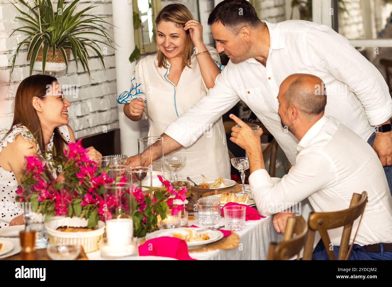 Gli amici sorridenti chiacchierano e ridono a una cena festiva. Due coppie sposate bevono cocktail e mangiano durante una riunione celebrativa. Giovani uomini e donne in grande stile Foto Stock