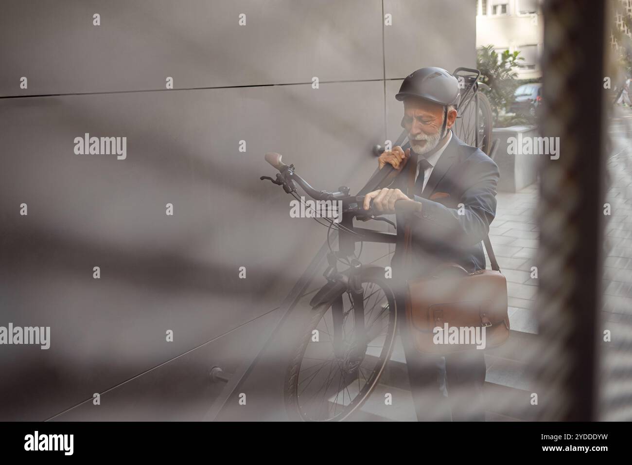 Senior manager con casco che porta la bicicletta sulla spalla mentre cammina su gradini visti attraverso la recinzione Foto Stock