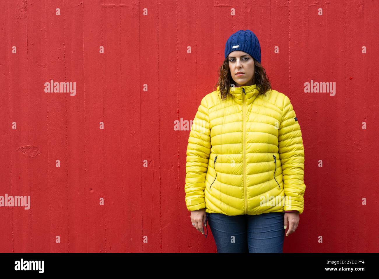 Ritratto di una donna bruna caucasica degli anni '30 che guarda seriamente la macchina fotografica, indossa un anorak giallo e un cappello di lana blu in posa con un pozzo rosso profondo della parete Foto Stock