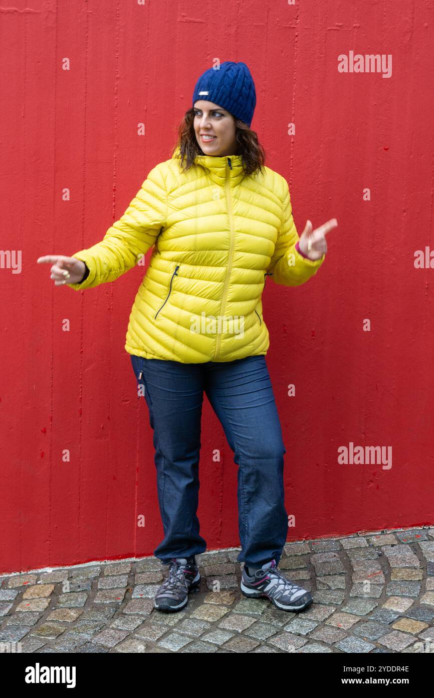 Una donna bruna con i suoi 30 anni che gestiva felicemente, indossando un anorak giallo e un cappello di lana blu con un muro rosso dietro di lei. Donna Street style Foto Stock