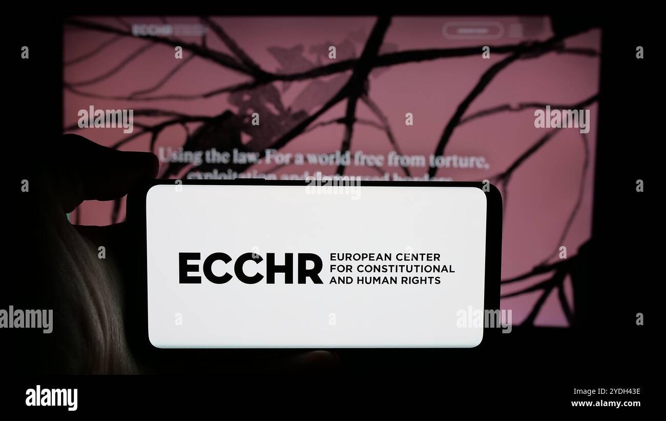 Persona che detiene un cellulare con il logo dell’organizzazione per i diritti umani ECCHR sullo schermo di fronte alla pagina web. Mettere a fuoco il display del telefono. Foto Stock