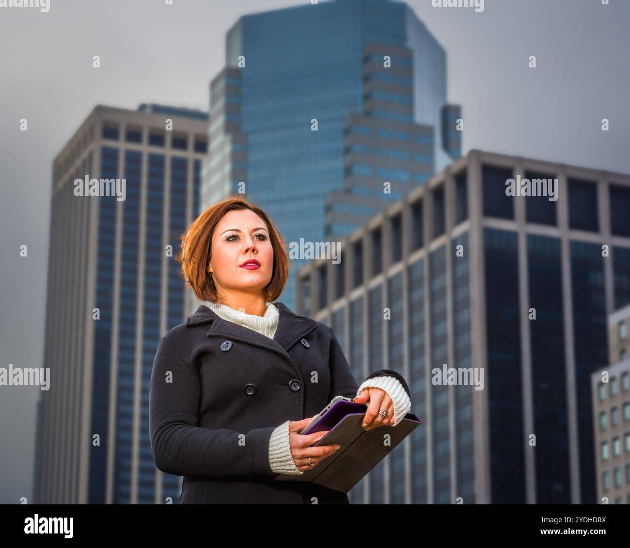 Una donna sta in piedi con sicurezza davanti a grattacieli torreggianti, tenendo un tablet in mano. Il cielo coperto e l'architettura moderna creano un vero e proprio mestiere Foto Stock