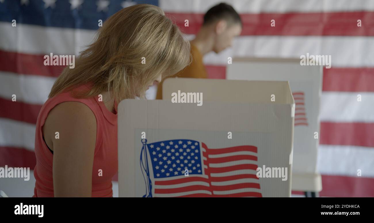 Profilo, medium shot, giovane donna dai capelli rossi che vota allo stand, guarda in basso durante la selezione dei candidati. Uomo asiatico e bandiera americana sullo sfondo Foto Stock