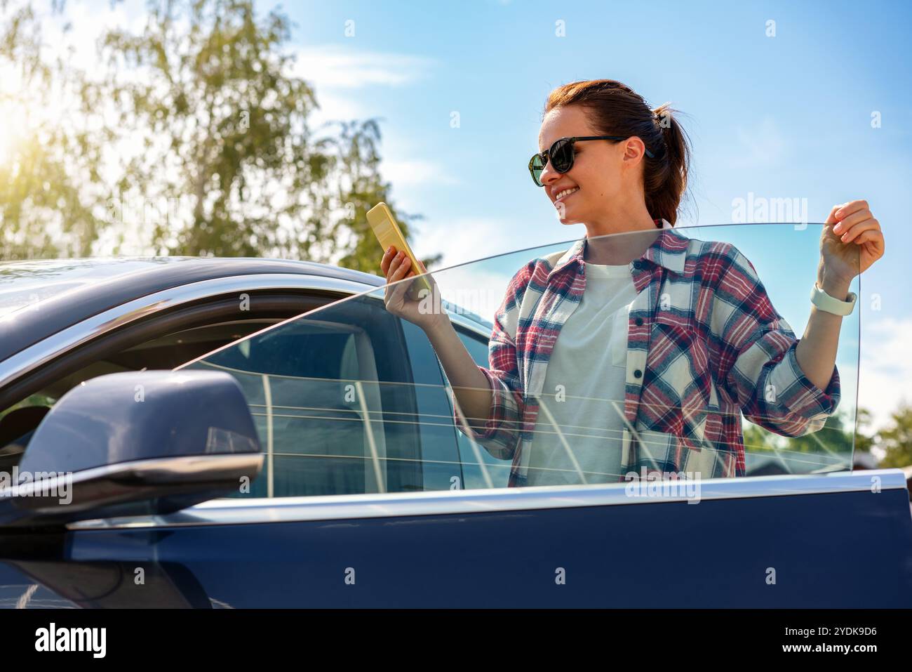 La bruna caucasica pilota utilizza lo smartphone mentre si trova accanto alla sua auto all'aperto. App mobile per car sharing. Servizio di noleggio auto. Foto Stock