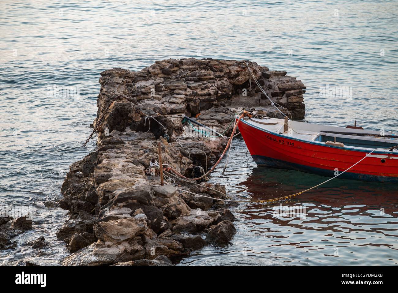 Una barca di legno rossa ormeggiata con corde a scogliere rocciose e scogli a Zante, in Grecia, la mattina presto. Il mare è calmo, la scena è tranquilla. Foto Stock