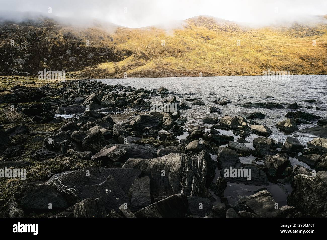 Un litorale roccioso si trova accanto a un tranquillo lago, con pendii erbosi che si innalzano sullo sfondo sotto un cielo nuvoloso. L'atmosfera tranquilla invita a riflettere Foto Stock