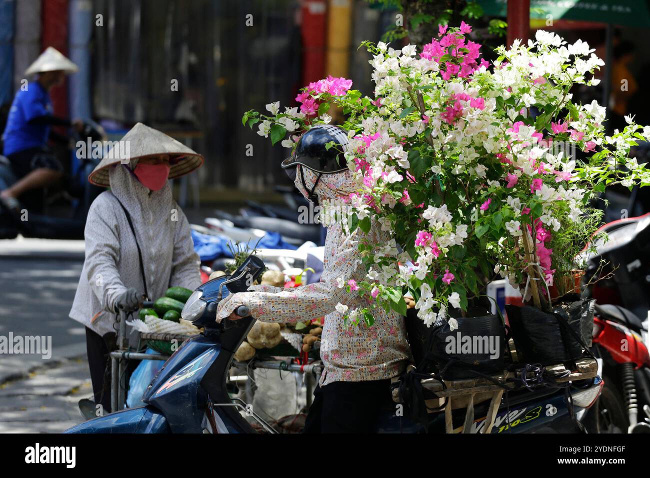 Hanoi, Vietnam - 23 luglio 2023: Il venditore di fiori vietnamita guida la sua moto attraverso una strada trafficata. Foto Stock